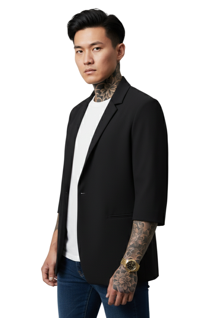 Man wearing a black blazer over a white shirt with tattoos on arms and neck, standing against a plain background.