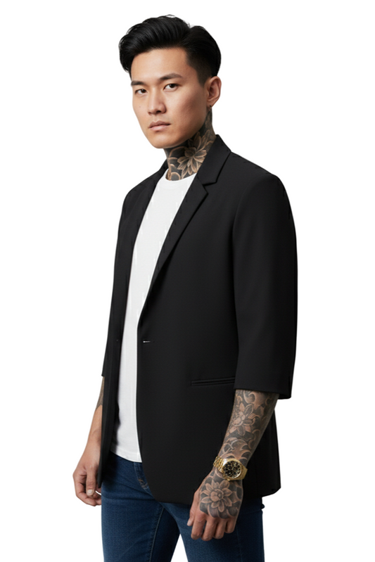 Man wearing a black blazer over a white shirt with tattoos on arms and neck, standing against a plain background.