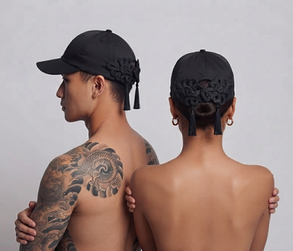 Two models shown from the back wearing matching black baseball caps with ornate black fabric embellishments and tassels, against a plain light gray studio background.