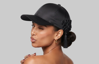 A woman shown in profile wearing a black satin baseball cap with a decorative black fabric flower and tassel on the side, against a clean white studio background.