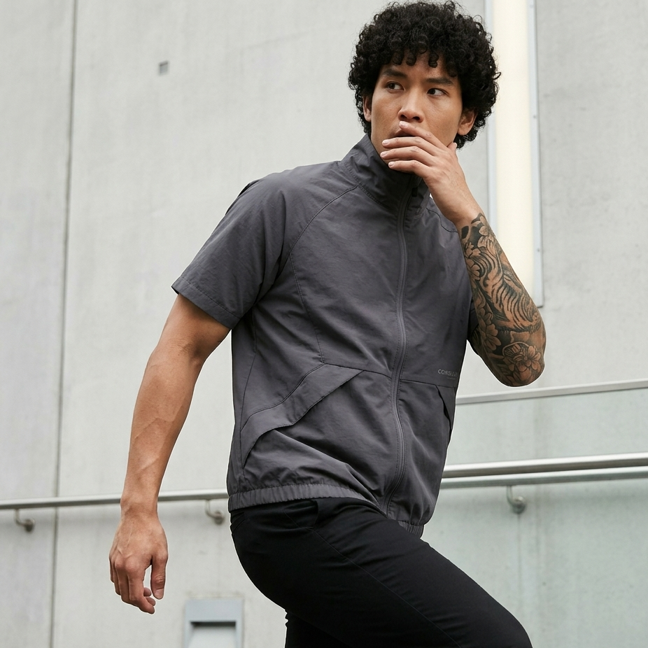 Man wearing a gray short-sleeve jacket and black pants against a light gray wall.