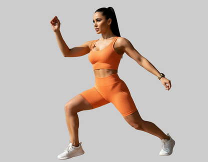 Woman in orange athletic wear performing a lunge in a room with wooden floor and gray walls.