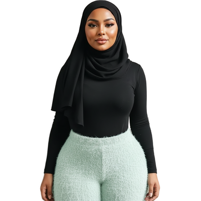 Woman wearing a black hijab, black long-sleeve top, and light gray pants against a beige wall.