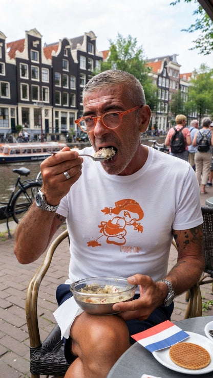 A man wearing orange-framed glasses and a white t-shirt with an orange cartoon graphic sits at an outdoor cafe table in Amsterdam, eating from a bowl while looking at the camera. The background shows canal houses and water.