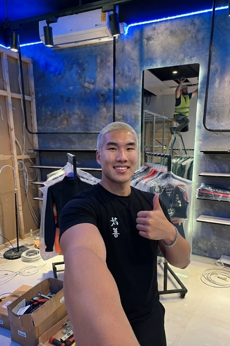 A man taking a selfie in a clothing store, wearing a black t-shirt with white Japanese characters on the chest and giving a thumbs-up. The background shows clothing racks, shelves, and a mirror reflecting a person on a ladder.