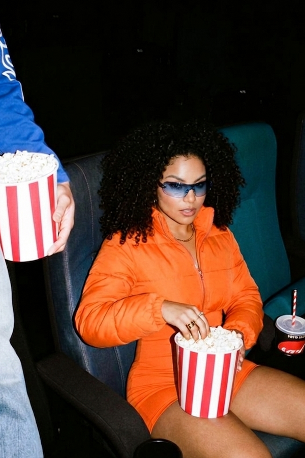 Woman in an orange jacket sitting in a movie theater with popcorn and a drink.