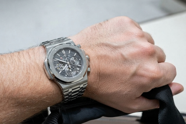 A close-up of a person's wrist wearing a silver-toned stainless steel chronograph watch with a dark dial and integrated metal bracelet, holding a dark strap in a real indoor setting.